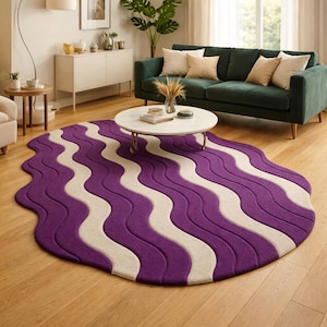 Può includere: Un tappeto a onde viola e crema in un soggiorno. Il tappeto, di forma ovale, presenta un motivo di onde viola e crema alternate. È posizionato su un pavimento in legno, con un divano verde e un tavolino bianco sullo sfondo.