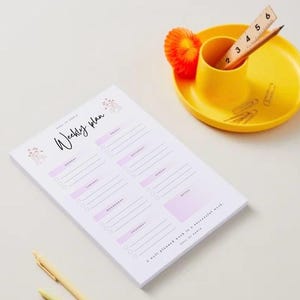 Könnte beinhalten: Ein weißer Wochenplan-Notizblock mit rosa und floralem Design, ein gelber Stifthalter und ein gelber Teller. Der Notizblock trägt die Aufschrift "Weekly Plan" und die Wochentage. Ein Holzlineal und ein Bleistift befinden sich im Halter.