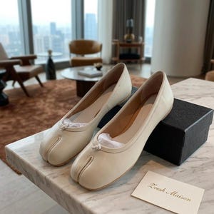 Può includere: Tazza in ceramica bianca con manico nero e la scritta "But First, Coffee" in nero. Lo sfondo sfocato suggerisce un'atmosfera accogliente.