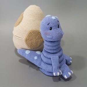 May include: A crocheted dinosaur toy in light blue with white spots, alongside a cream-coloured egg with brown patches. The dinosaur has a long neck and a friendly face, suitable for a child's bedroom.