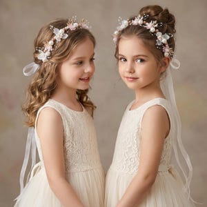 May include: Two floral headbands featuring white and pink petals, small pearls, and gold-toned accents. Each headband includes long, sheer white ribbon ties that drape down. The headbands are worn as hair accessories with cream-colored lace dresses.