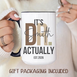 May include: White ceramic mug with black handle and rim, featuring the text "IT'S DR. Smith ACTUALLY EST.2026" in brown and black lettering. The mug is held by a person wearing a white sweater.