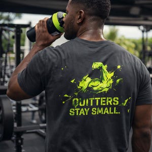 Puede incluir: Camiseta gris oscuro con un diseño gráfico verde neón de un brazo y el texto "QUITTERS STAY SMALL". Una persona lleva la camiseta y bebe de una botella de agua negra y verde en un gimnasio.