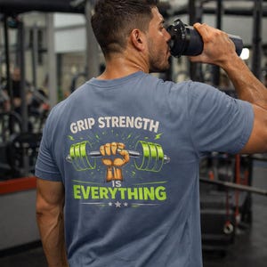 May include: A blue t-shirt with the text "GRIP STRENGTH IS EVERYTHING" and a graphic of a weight bar held by a fist. The weight plates are green. The shirt is being worn in a gym setting.