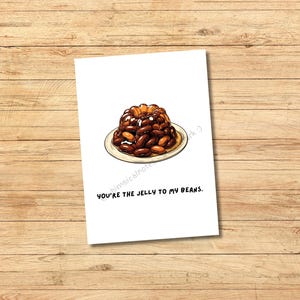 Könnte beinhalten: Eine weiße Grußkarte mit einer Cartoon-Illustration eines Wackelpudding-Forms mit Bohnen auf einem Teller. Der Text "You're the jelly to my beans" ist unter dem Bild aufgedruckt. Die Karte liegt auf einer Holzoberfläche.