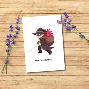 May include: A white card with a cartoon illustration of a thief carrying a sack filled with a heart. The text "YOU STOLE MY HEART." is below the illustration. Lavender sprigs are on a wooden surface.