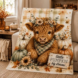 May include: A soft throw blanket featuring a fluffy highland cow wearing a floral crown and a plaid bandana, surrounded by pumpkins and autumn leaves. The blanket includes a wooden sign graphic with the text, little PUMPKIN big dreams.