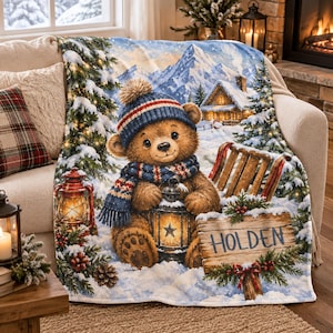 Könnte beinhalten: Eine weiche Wohndecke mit einem braunen Teddybären, der eine blau-rote Strickmütze und einen Schal trägt und eine Laterne hält. Das Motiv zeigt eine verschneite Berglandschaft, eine Hütte, Tannenbäume und ein Holzschild mit dem Namen HOLDEN.