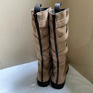 May include: A pair of tan suede knee-high boots with black zippers and straps. The boots have a rounded toe and a black sole. The zippers run down the back of the boots, and the straps are decorative.