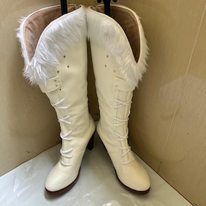Op de afbeelding: Ivoor kniehoge laarzen met een vetersluiting en een kleine hak. De bovenkant van de laarzen is afgezet met witte imitatiebont. De laarzen worden gepresenteerd tegen een neutrale achtergrond. De hakhoogte is ongeveer 7,6 cm.