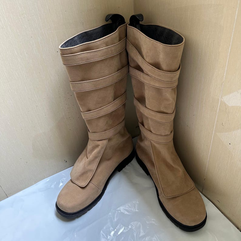 May include: A pair of tan knee-high boots with multiple horizontal straps and a rounded toe. The boots have a suede-like texture and a black sole. The interior of the boot is black. The boots are standing upright.