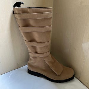 May include: A tan suede boot with a black sole. The boot features multiple horizontal straps and a flap closure on the side. A black boot shaper is inserted in the boot.