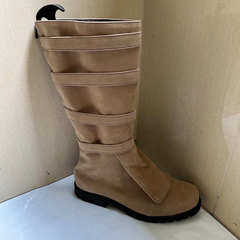 May include: A tan suede boot with a black sole. The boot features multiple horizontal straps and a flap closure on the side. A black boot shaper is inserted in the boot.