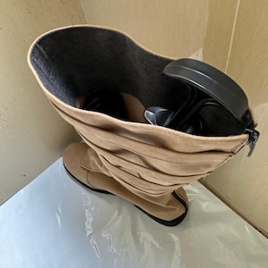 May include: Tan suede knee-high boot with a black sole and a zipper closure. The boot has a ruched design and a dark gray interior. A black shoe tree is inside the boot.