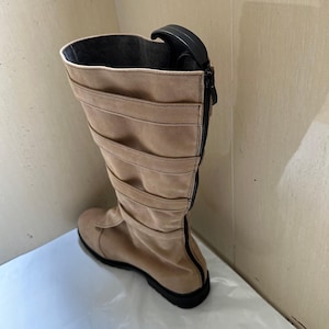 May include: A tan suede-like boot with a black zipper and sole. The boot features horizontal stitched accents and a black handle at the top. The interior is lined with a darker fabric. This footwear is suitable for various occasions.