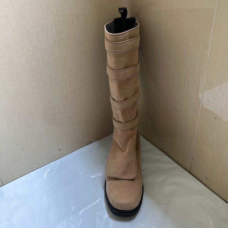 May include: A tan suede knee-high boot with multiple horizontal straps and a black sole. The boot has a rounded toe and a pull tab at the top. The boot is made of a soft, suede-like material.