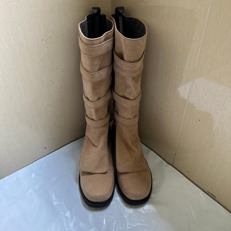 May include: A pair of tall, tan suede boots with multiple straps and a rounded toe. The boots have a black sole and a contrasting cream-colored trim around the top opening. The boots are standing upright.