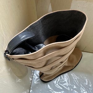 May include: Tan suede knee-high boots with a stacked heel. The boots have a rounded toe and a series of horizontal pleats. Inside the boot, there are black high heels. The interior of the boot is lined with gray fabric.