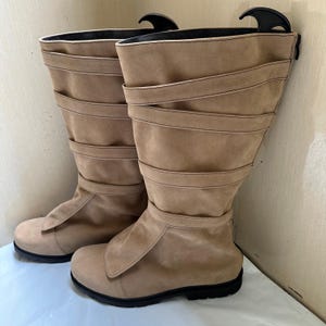 May include: A pair of tan suede-like boots. The boots have a knee-high design with multiple horizontal straps and a flap over the foot. The soles are black. The boots are displayed on a white surface.