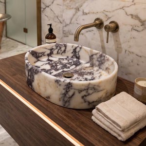May include: An oval marble vessel sink with white and deep purple veining sits atop a dark wood floating vanity. A wall-mounted brass faucet is positioned above the sink. A stack of folded white towels and a small candle sit on the vanity surface.