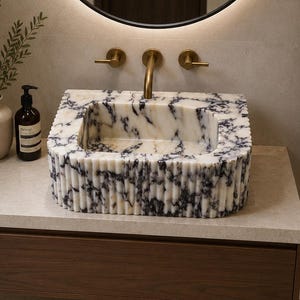 May include: A rectangular marble sink with a white and dark blue veined pattern and a fluted exterior design. The sink is mounted on a light-colored stone countertop above a dark wood floating vanity drawer, featuring brass faucet fixtures.