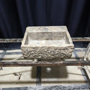 K&ouml;nnte beinhalten: Ein rechteckiges, verwittertes Steinwaschbecken mit einer rauen, strukturierten Au&szlig;enseite. Das Waschbecken hat ein zentrales Abflussloch und steht auf einem Metallrahmen. Der Hintergrund ist ein dunkler Vorhang.