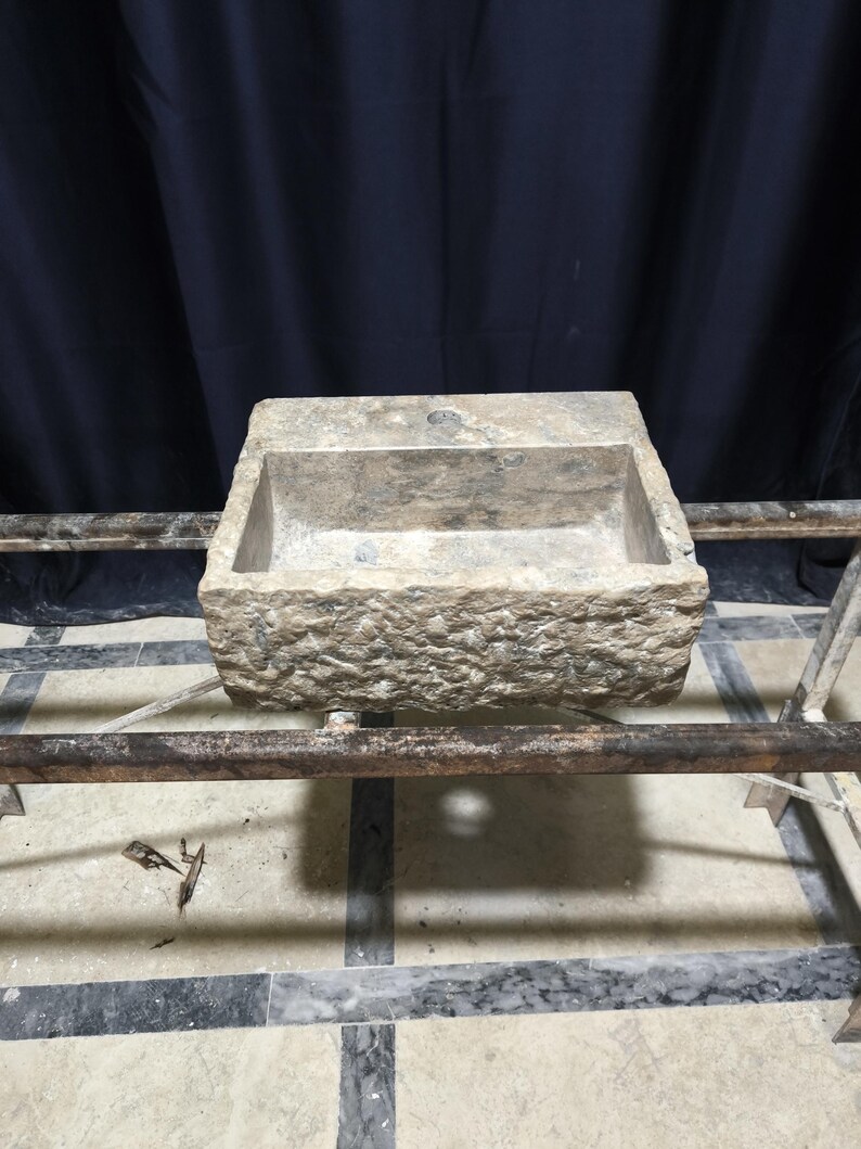 K&ouml;nnte beinhalten: Ein rechteckiges, verwittertes Steinwaschbecken mit einer rauen, strukturierten Au&szlig;enseite. Das Waschbecken hat ein zentrales Abflussloch und steht auf einem Metallrahmen. Der Hintergrund ist ein dunkler Vorhang.