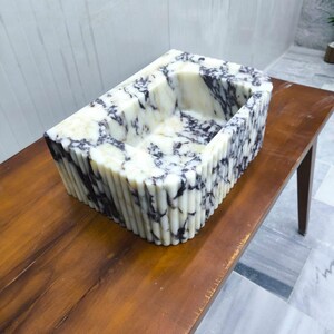 May include: A rectangular stone sink featuring a white base with prominent dark purple and black veining throughout. The exterior of the basin is designed with a vertical fluted texture, while the interior maintains a smooth, polished finish.