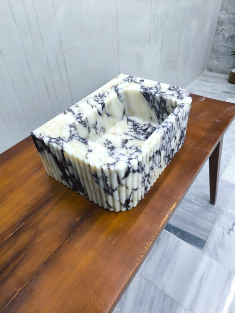May include: A rectangular stone sink featuring a white base with prominent dark purple and black veining throughout. The exterior of the basin is designed with a vertical fluted texture, while the interior maintains a smooth, polished finish.
