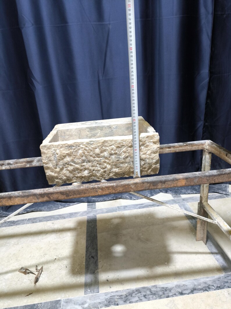K&ouml;nnte beinhalten: Ein rechteckiger, hellfarbener Steinpflanzer mit rauer, strukturierter Oberfl&auml;che. Ein Metallmessband zeigt die H&ouml;he des Pflanzgef&auml;&szlig;es. Der Pflanzer steht auf einem Metallrahmen.