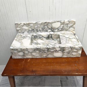 May include: A rectangular marble sink with a built-in backsplash. The sink basin is a recessed square. The marble has a white base with gray veining. The sink is on a brown wooden table.
