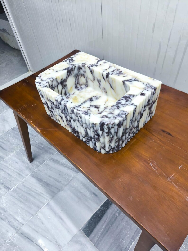 May include: A rectangular stone sink featuring a white base with prominent dark purple and black veining. The exterior of the basin is constructed with a series of vertical fluted columns, creating a textured, ribbed appearance around the entire perimeter.