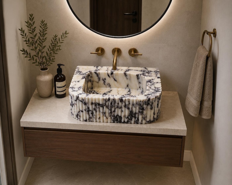 May include: A rectangular marble sink with a white and dark blue veined pattern and a fluted exterior design. The sink is mounted on a light-colored stone countertop above a dark wood floating vanity drawer, featuring brass faucet fixtures.