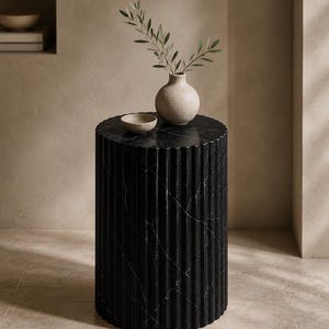 Pode incluir: Uma mesa de apoio cilíndrica em mármore preto com uma superfície exterior estriada verticalmente. Sobre a superfície plana encontram-se uma pequena taça de cerâmica de cor clara e um vaso esférico a condizer com um ramo de oliveira.