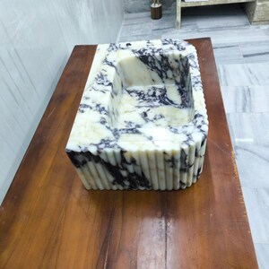 May include: A rectangular bathroom sink carved from white marble with prominent dark purple veining. The exterior features a vertical fluted design, while the interior basin is smooth and deep, showcasing the natural stone pattern throughout.