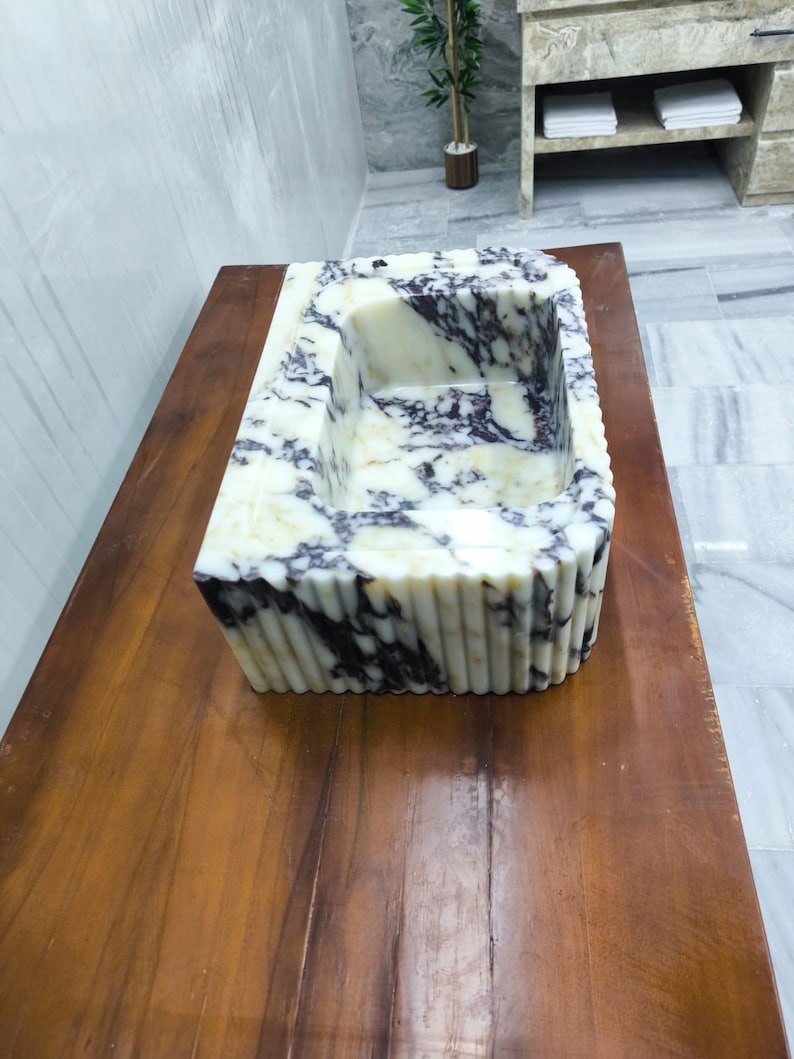 May include: A rectangular bathroom sink carved from white marble with prominent dark purple veining. The exterior features a vertical fluted design, while the interior basin is smooth and deep, showcasing the natural stone pattern throughout.