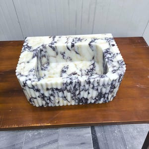 May include: A rectangular stone sink featuring a white base with prominent dark purple and black veining. The exterior walls of the basin display a vertical fluted or ribbed texture, while the interior basin is smooth with a deep rectangular cavity.