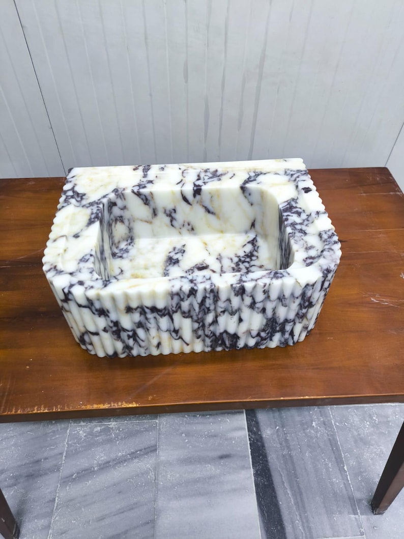 May include: A rectangular stone sink featuring a white base with prominent dark purple and black veining. The exterior walls of the basin display a vertical fluted or ribbed texture, while the interior basin is smooth with a deep rectangular cavity.