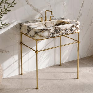Puede incluir: Un lavabo de mármol con grifos dorados y un soporte de metal dorado. La pila del lavabo es blanca con vetas marrón oscuro. El grifo y los tiradores son dorados. El soporte tiene un diseño geométrico.