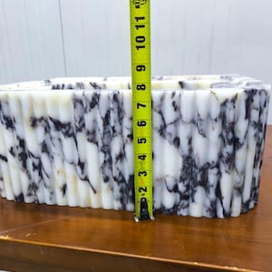 May include: A rectangular white and dark purple marble vessel with a fluted, ribbed exterior texture. A yellow measuring tape is positioned vertically against the front, indicating a height of approximately 8.5 inches from the base to the top rim.