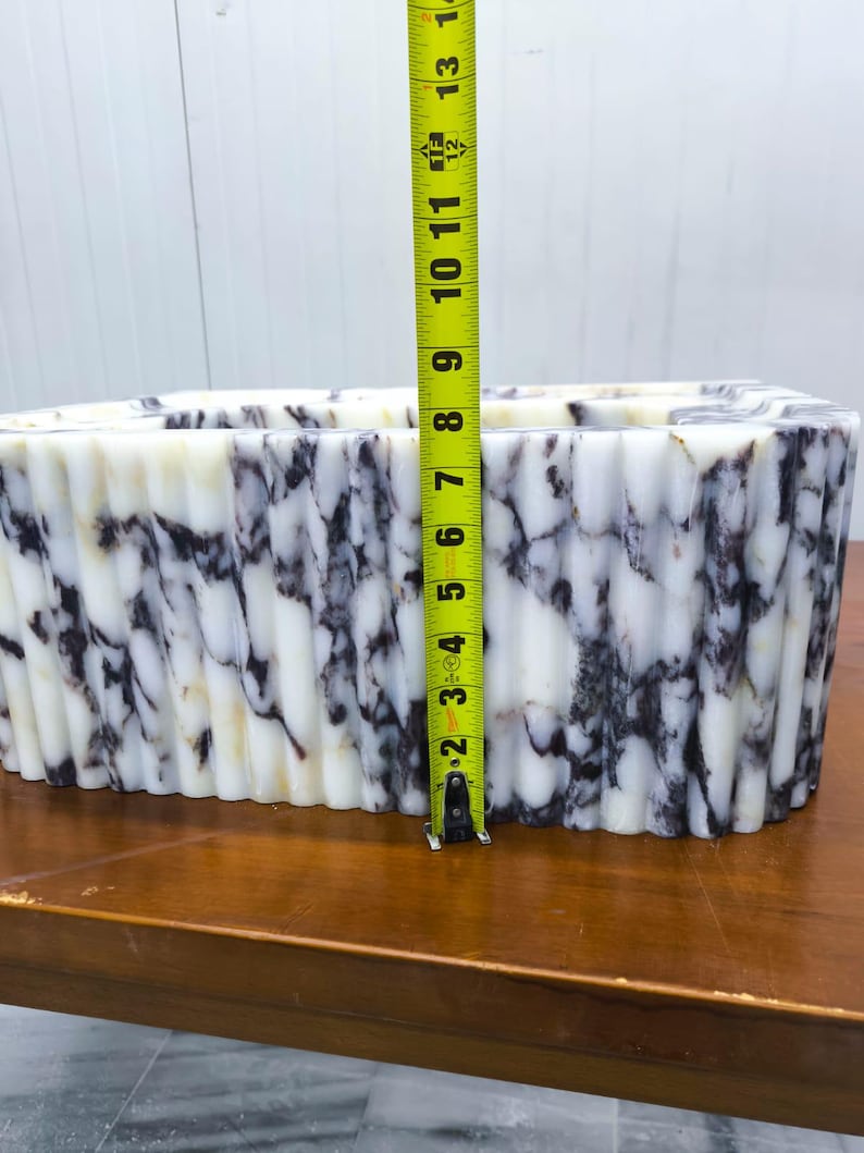 May include: A rectangular white and dark purple marble vessel with a fluted, ribbed exterior texture. A yellow measuring tape is positioned vertically against the front, indicating a height of approximately 8.5 inches from the base to the top rim.