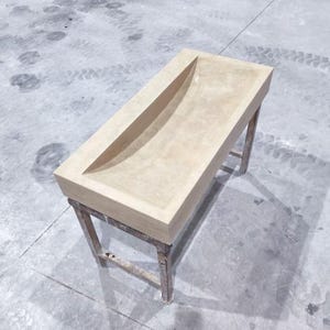 Peut inclure: Un évier rectangulaire en béton beige clair avec un long bassin incurvé. L'évier est soutenu par un cadre métallique vieilli. Le design est minimaliste et moderne, adapté à une salle de bain ou à un espace extérieur.