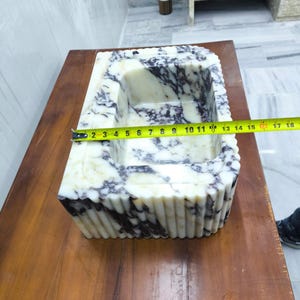 May include: A rectangular marble sink featuring a white and deep purple veined pattern with a fluted exterior design. A yellow measuring tape is placed across the basin, indicating a width of approximately 18 inches from edge to edge.