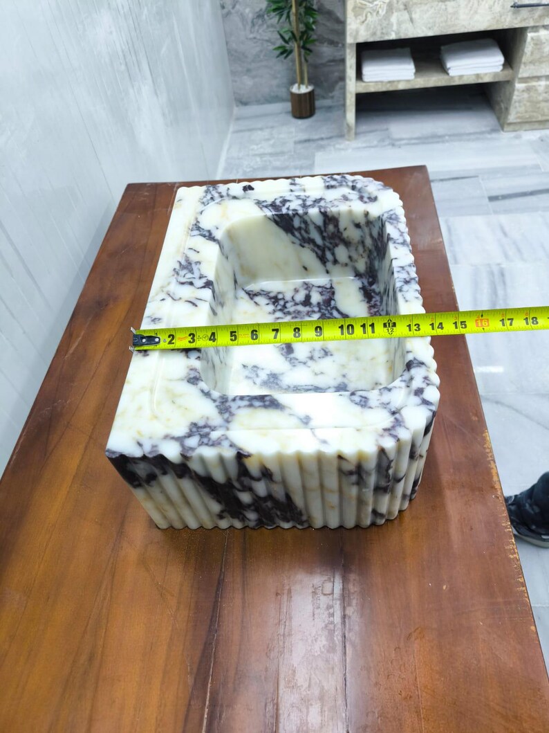 May include: A rectangular marble sink featuring a white and deep purple veined pattern with a fluted exterior design. A yellow measuring tape is placed across the basin, indicating a width of approximately 18 inches from edge to edge.