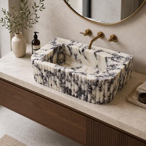 May include: A rectangular bathroom sink crafted from white marble with prominent black veining. The exterior features a vertical fluted texture, and the basin is integrated into a countertop. It includes a brass faucet with two handles.