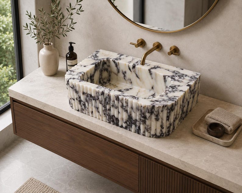 May include: A rectangular bathroom sink crafted from white marble with prominent black veining. The exterior features a vertical fluted texture, and the basin is integrated into a countertop. It includes a brass faucet with two handles.