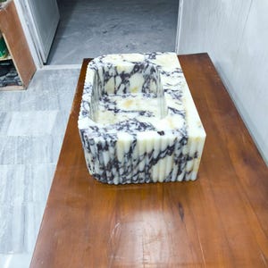 May include: A rectangular stone sink featuring a polished white surface with deep purple and black veining. The exterior of the basin displays a vertical fluted or ribbed texture, while the interior basin is smooth with a consistent marble pattern.