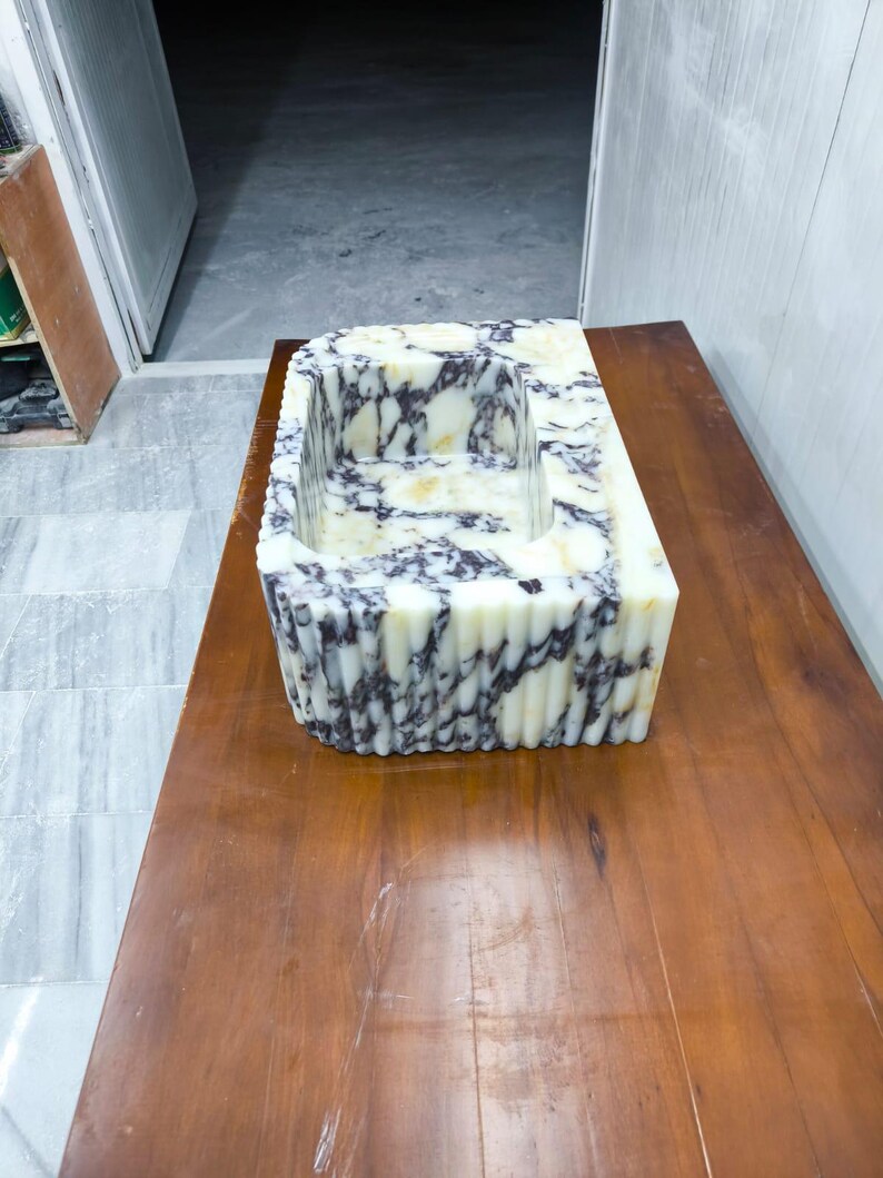 May include: A rectangular stone sink featuring a polished white surface with deep purple and black veining. The exterior of the basin displays a vertical fluted or ribbed texture, while the interior basin is smooth with a consistent marble pattern.