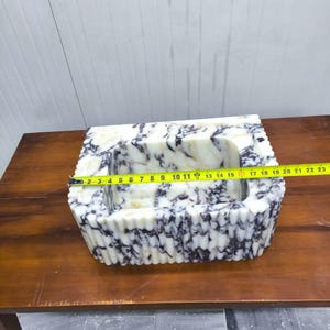 May include: A rectangular white and purple marble sink with a fluted exterior design. A yellow measuring tape is stretched across the top, indicating a width of approximately 19 inches from the outer edges of the stone basin structure.