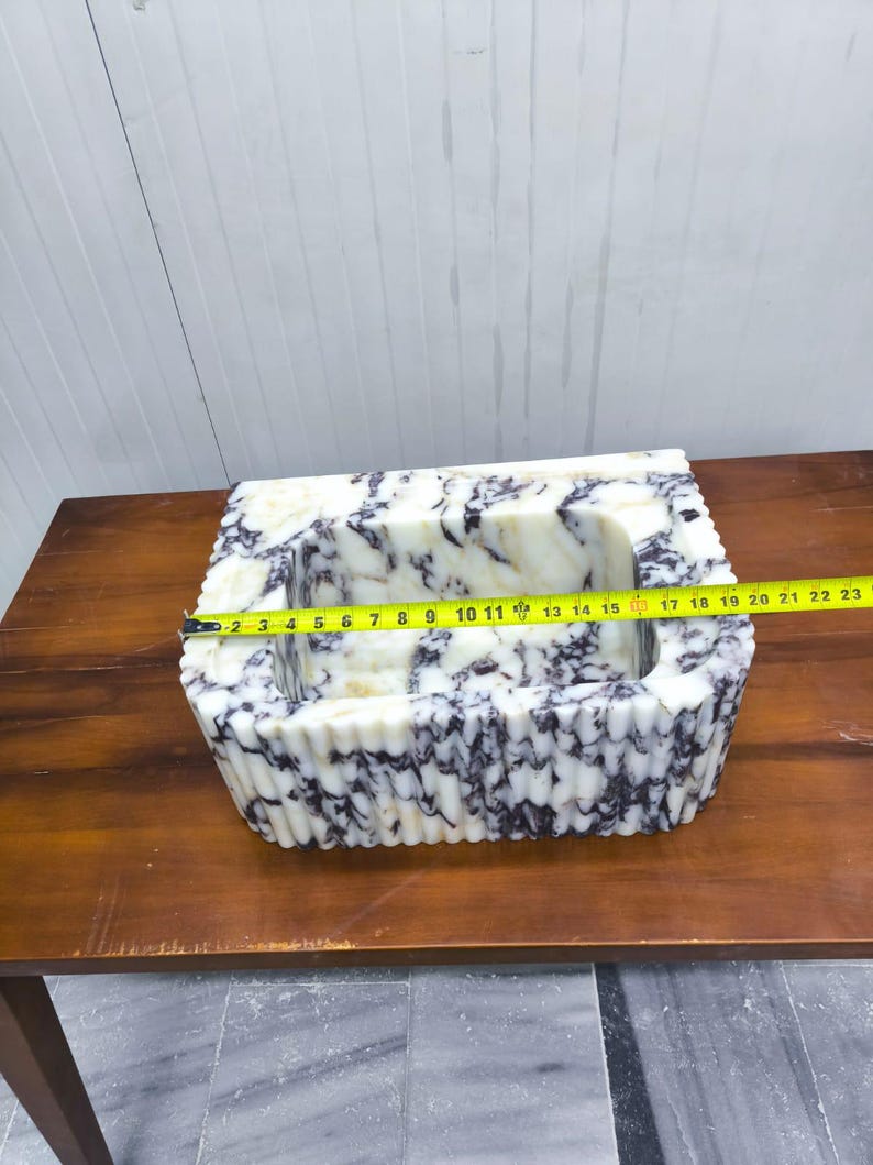 May include: A rectangular white and purple marble sink with a fluted exterior design. A yellow measuring tape is stretched across the top, indicating a width of approximately 19 inches from the outer edges of the stone basin structure.